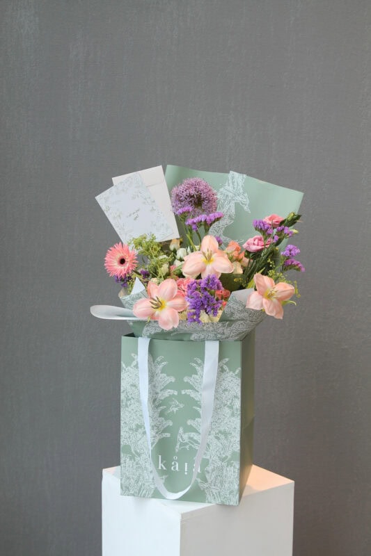 Elegant floral arrangement with pink lilies, purple alliums, and gerbera daisies in a decorative gift bag.