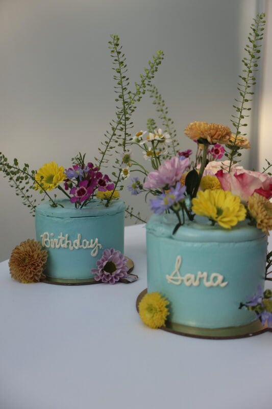 Two beautifully decorated birthday cakes with pastel blue frosting, adorned with fresh colorful flowers and greenery, perfect for celebrations and special occasions.