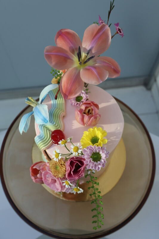Elegant pink floral cake decorated with tulips, daisies, and greenery, perfect for celebrations and special occasions.