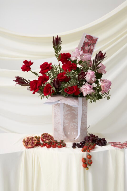 Elegant floral arrangement with red and pink roses, complemented by dark foliage, arranged in a white gift box on a white draped background.