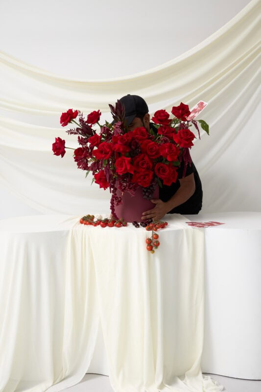 Elegant red rose bouquet in a pink vase with lush greenery, displayed on a white draped background for a sophisticated floral arrangement.