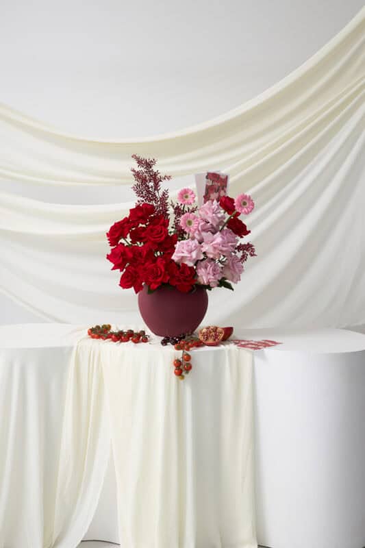 Beautiful floral display featuring vibrant red and soft pink flowers arranged in a deep burgundy vase, set against a flowing white fabric backdrop for a sophisticated look.