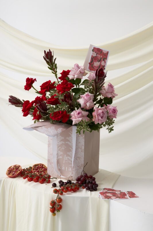 Elegant bouquet of red and pink roses arranged in a white vase, complemented by fresh grapes and decorative cards, perfect for floral gifting and special occasions.