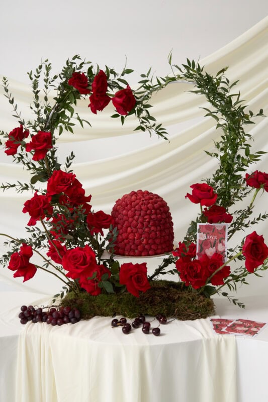 Beautiful floral arrangement featuring vibrant red roses forming a heart shape, accented with lush green foliage, set against a soft white backdrop. Perfect for romantic occasions and special celebrat.