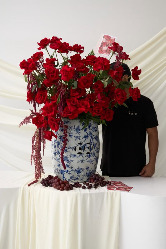 Elegant red floral arrangement featuring roses and carnations in a blue and white porcelain vase, displayed on a white table with a soft draped background.