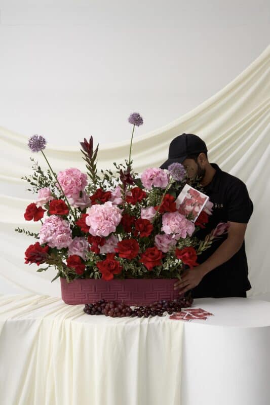 A man arranging a stunning flower bouquet featuring pink hydrangeas, red roses, and purple alliums in a pink basket, set against a soft white backdrop.