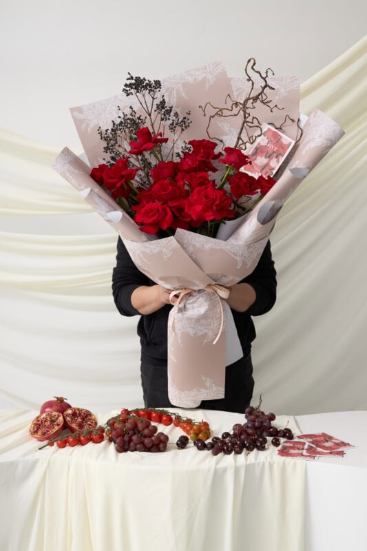 Elegant red rose bouquet with decorative foliage, wrapped in soft pink paper, held by a person at a floral event.