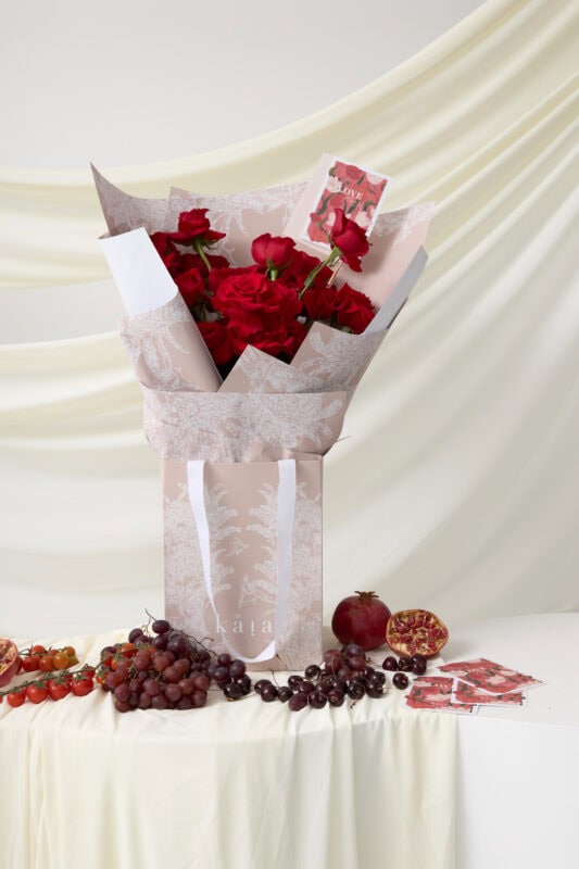 Elegant bouquet of red roses wrapped in decorative paper, displayed on a white table with grapes and pomegranate, perfect for special occasions.