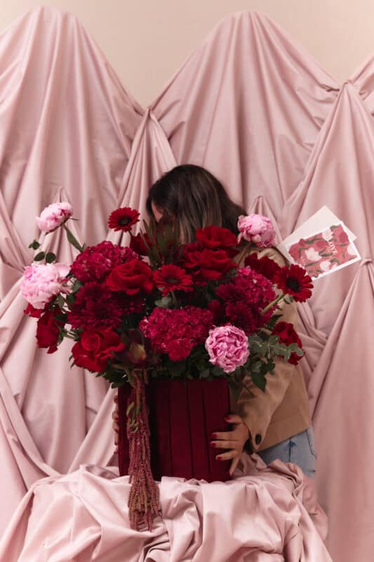 Woman holding a large bouquet of red and pink flowers in front of draped pink fabric backdrop, emphasizing floral arrangement and romantic theme.