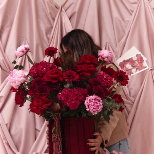 Woman holding a large bouquet of red and pink flowers in front of draped pink fabric backdrop, emphasizing floral arrangement and romantic theme.