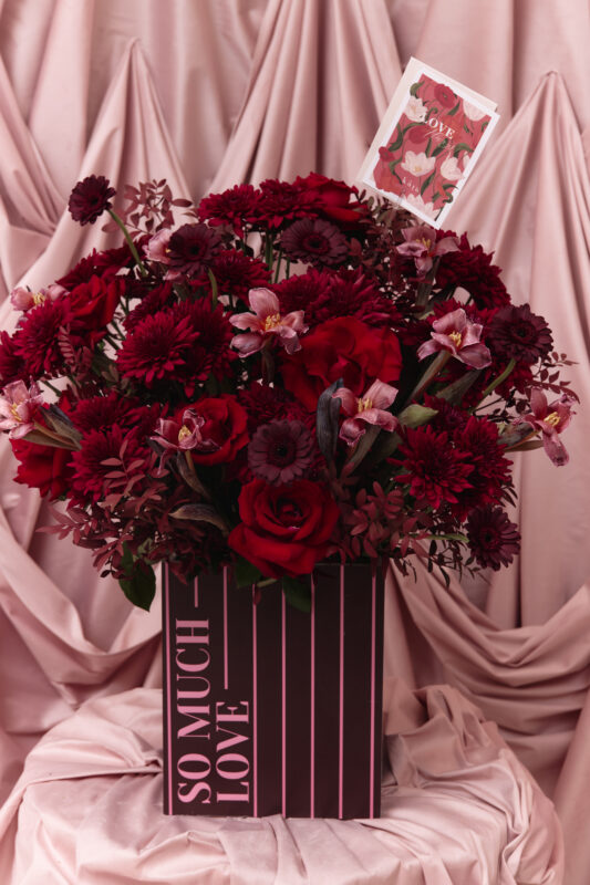 Beautiful Valentine's Day flower arrangement with roses, gerbera daisies, and orchids in a pink striped box. Perfect for expressing love and affection.