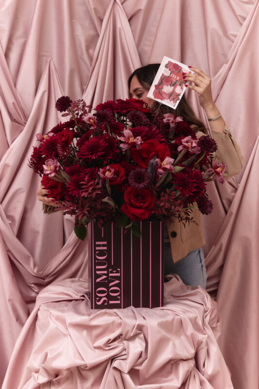 A woman holding a large bouquet of vibrant red flowers, including roses and gerbera daisies, in front of a soft pink draped fabric backdrop. She is also holding a card with a love theme, emphasizing r.