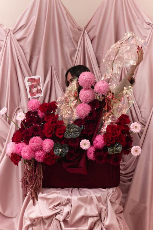 Elegant floral display featuring pink and red roses with dark green foliage, set against soft pink draped fabric for a romantic and dreamy aesthetic.