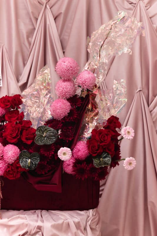 Beautiful pink and red floral bouquet with roses and pom-poms, set against a soft pink draped fabric background. Perfect for romantic or special occasions.