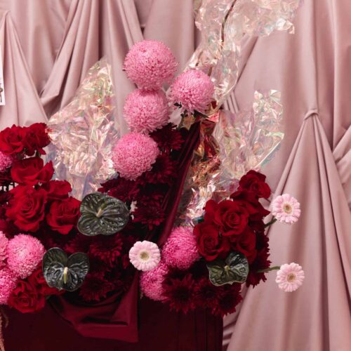 Beautiful pink and red floral bouquet with roses and pom-poms, set against a soft pink draped fabric background. Perfect for romantic or special occasions.