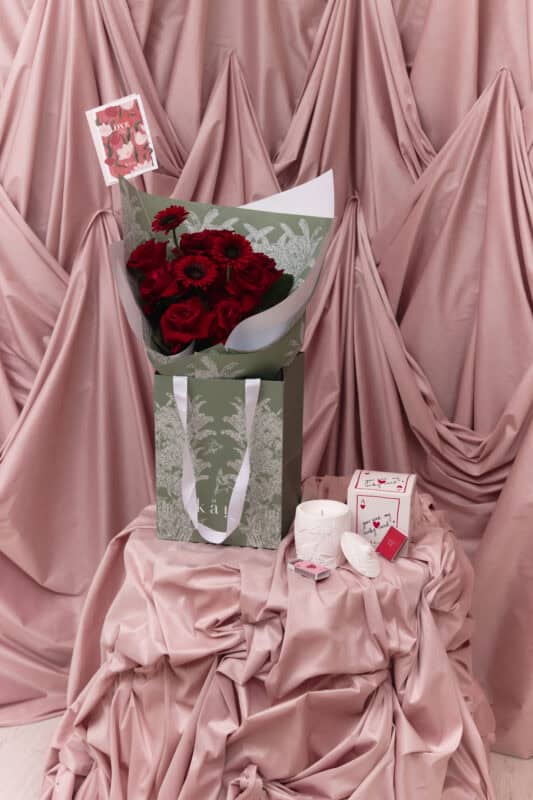 Elegant bouquet of deep red roses and gerbera daisies wrapped in white and green paper, set against soft pink draped fabric, symbolizing eternal love and romance.