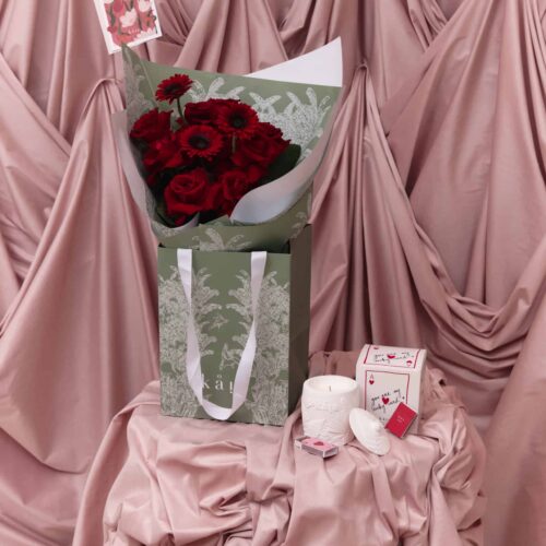 Elegant bouquet of deep red roses and gerbera daisies wrapped in white and green paper, set against soft pink draped fabric, symbolizing eternal love and romance.