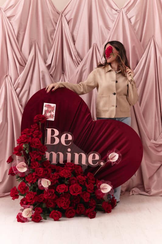 An elegant woman holding a large heart-shaped floral display with red roses and pink calla lilies, set against a soft pink draped backdrop for a romantic Valentine's Day theme.