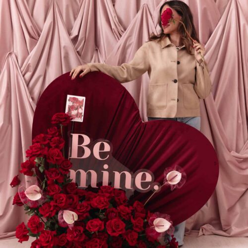 An elegant woman holding a large heart-shaped floral display with red roses and pink calla lilies, set against a soft pink draped backdrop for a romantic Valentine's Day theme.