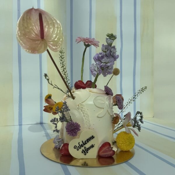 Elegant celebration cake decorated with fresh flowers, strawberries, and a "Welcome Home" heart-shaped topper, perfect for special occasions.