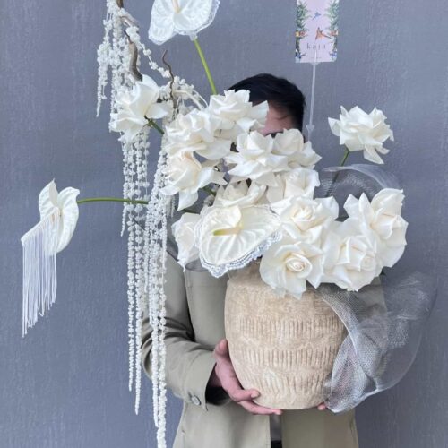 Beautiful white floral arrangement with an assortment of roses, anthuriums, and cascading flowers in a large beige vase, set against a gray background.