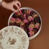 A hand reaching into a round box filled with assorted flower-shaped cookies in pink, brown, and beige, with a decorative lid featuring floral and animal illustrations.