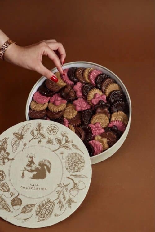 A hand reaching into a round box filled with assorted flower-shaped cookies in pink, brown, and beige, with a decorative lid featuring floral and animal illustrations.