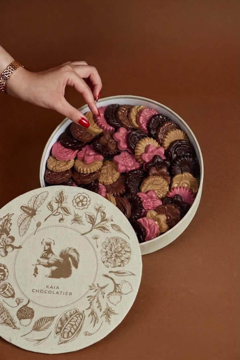 A hand reaching into a round box filled with assorted flower-shaped cookies in pink, brown, and beige, with a decorative lid featuring floral and animal illustrations.
