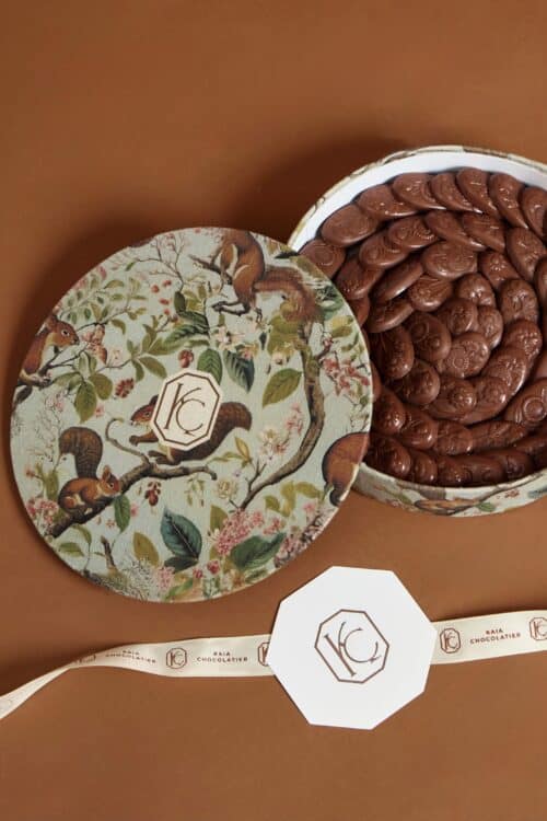 Elegant round gift box filled with chocolate ovals, featuring a nature-inspired design with squirrels and leaves, perfect for gifting.
