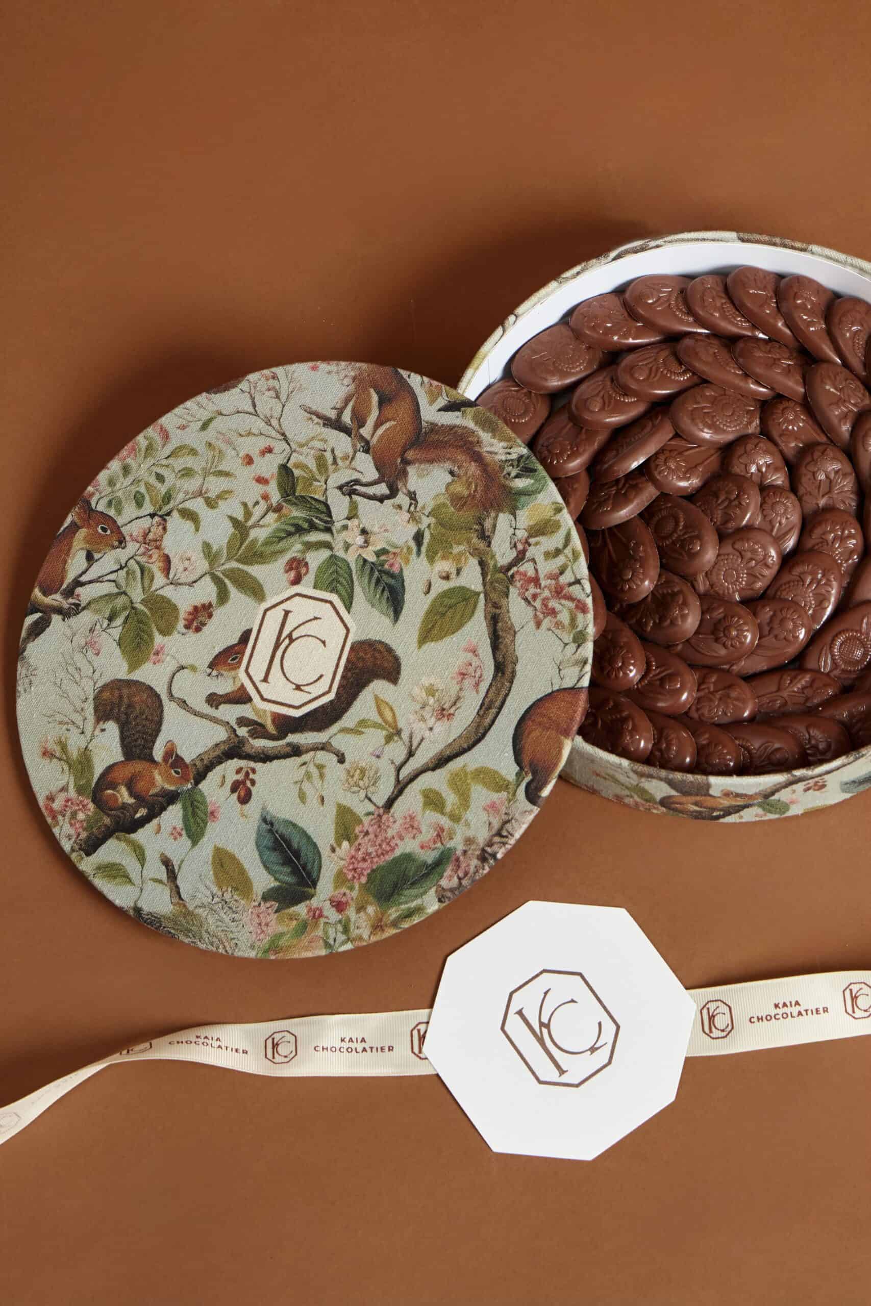 Elegant round gift box filled with chocolate ovals, featuring a nature-inspired design with squirrels and leaves, perfect for gifting.