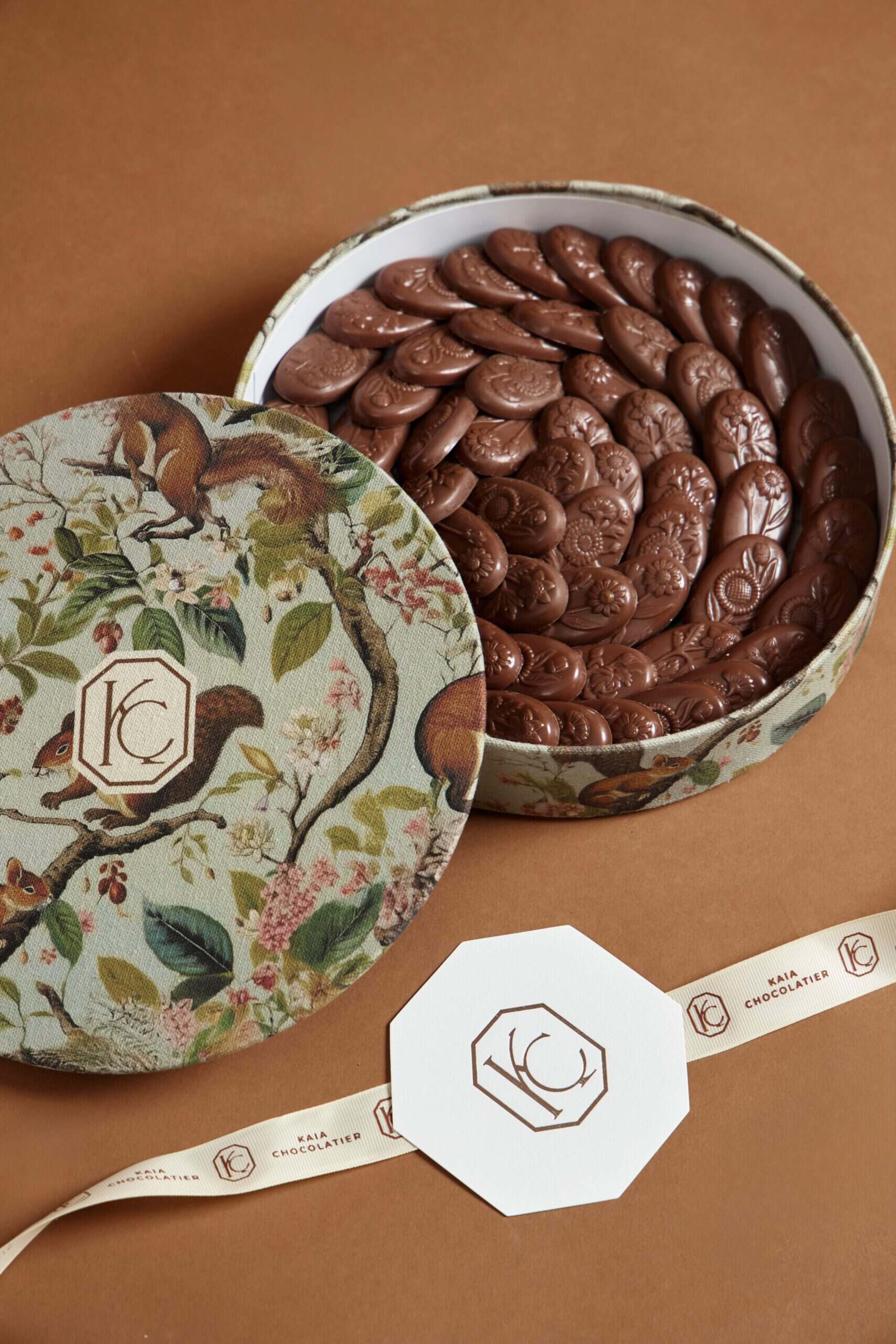 Elegant round gift box filled with glossy chocolate ovals, decorated with a floral and animal print lid, perfect for gifting or indulgence.