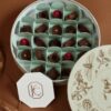 Assorted chocolates in a round box with decorative lid, featuring various shapes and toppings, on a brown surface.