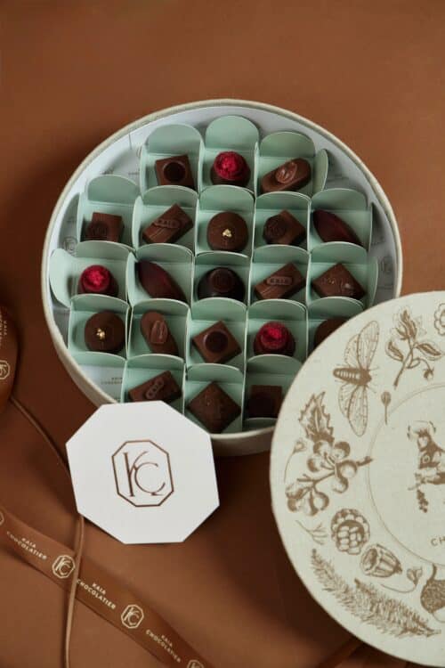 Assorted chocolates in a round box with decorative lid, featuring various shapes and toppings, on a brown surface.