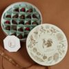Elegant round chocolate box with floral and squirrel illustrations, filled with assorted chocolates and red berries, on a brown background.