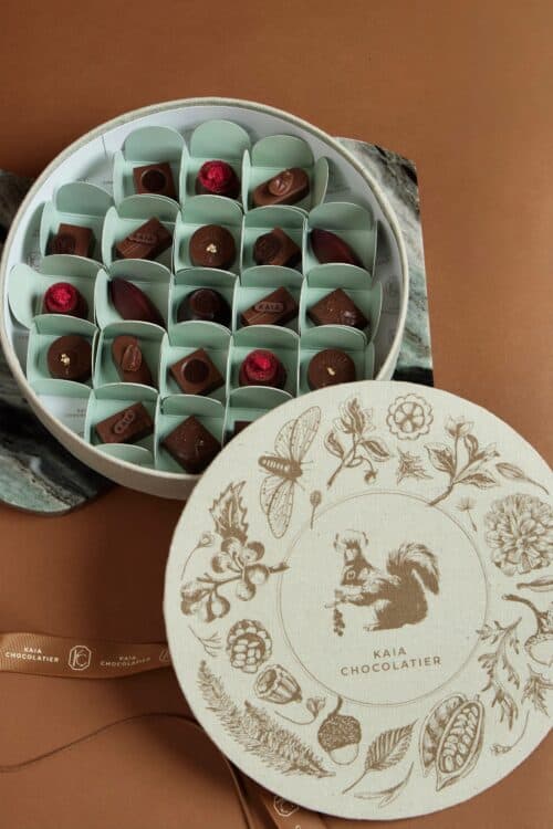 Elegant round box of assorted chocolates with floral and nature-inspired illustrations on the lid, featuring a squirrel and botanical elements, perfect for gift-giving.