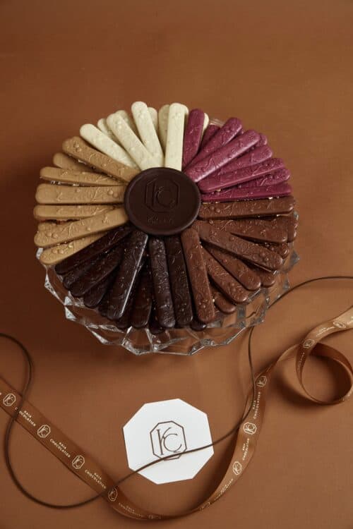 Assorted chocolate bird sticks arranged in a circular pattern on a glass cake stand, featuring various flavors and colors for dessert decoration.