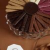 Assorted chocolate bird sticks in various flavors and colors arranged in a circular pattern on a glass plate, with a Kaia Flowers logo in the foreground.
