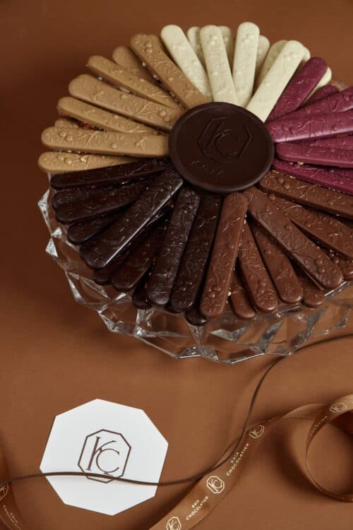 Assorted chocolate bird sticks in various flavors and colors arranged in a circular pattern on a glass plate, with a Kaia Flowers logo in the foreground.