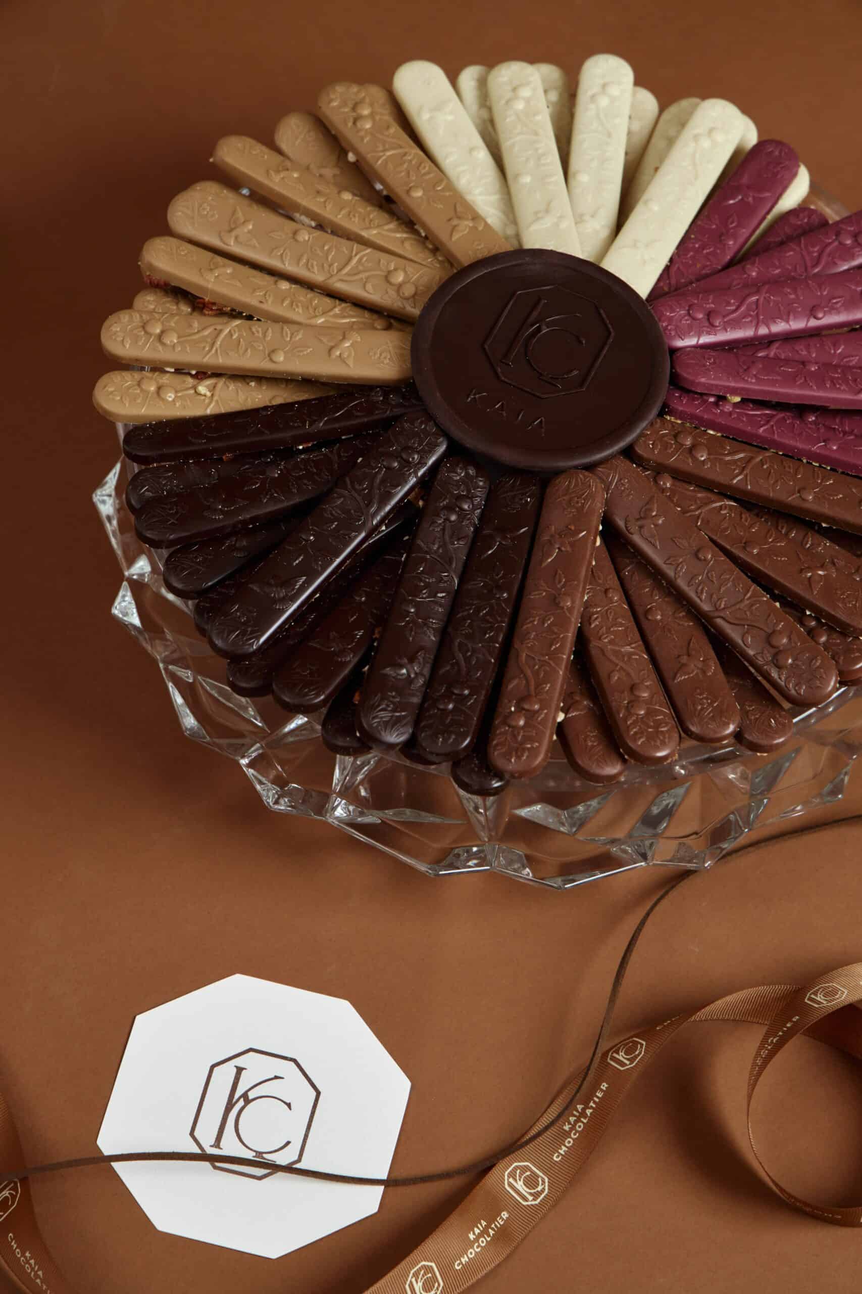 Assorted chocolate bird sticks in various flavors and colors arranged in a circular pattern on a glass plate, with a Kaia Flowers logo in the foreground.